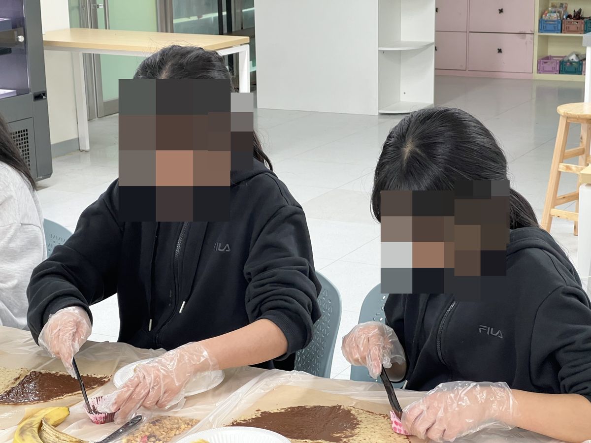 나란히 앉아 크레페에 초코 스프레드를 바르는 손과 분홍 종이컵, 검은 플라스틱 나이프