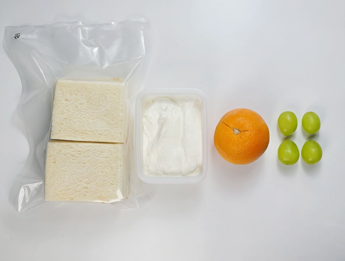 비닐에 든 식빵 두 장, 흰 크림 용기, 오렌지 한 개, 포도 알이 나란히 놓인 키트 식재료