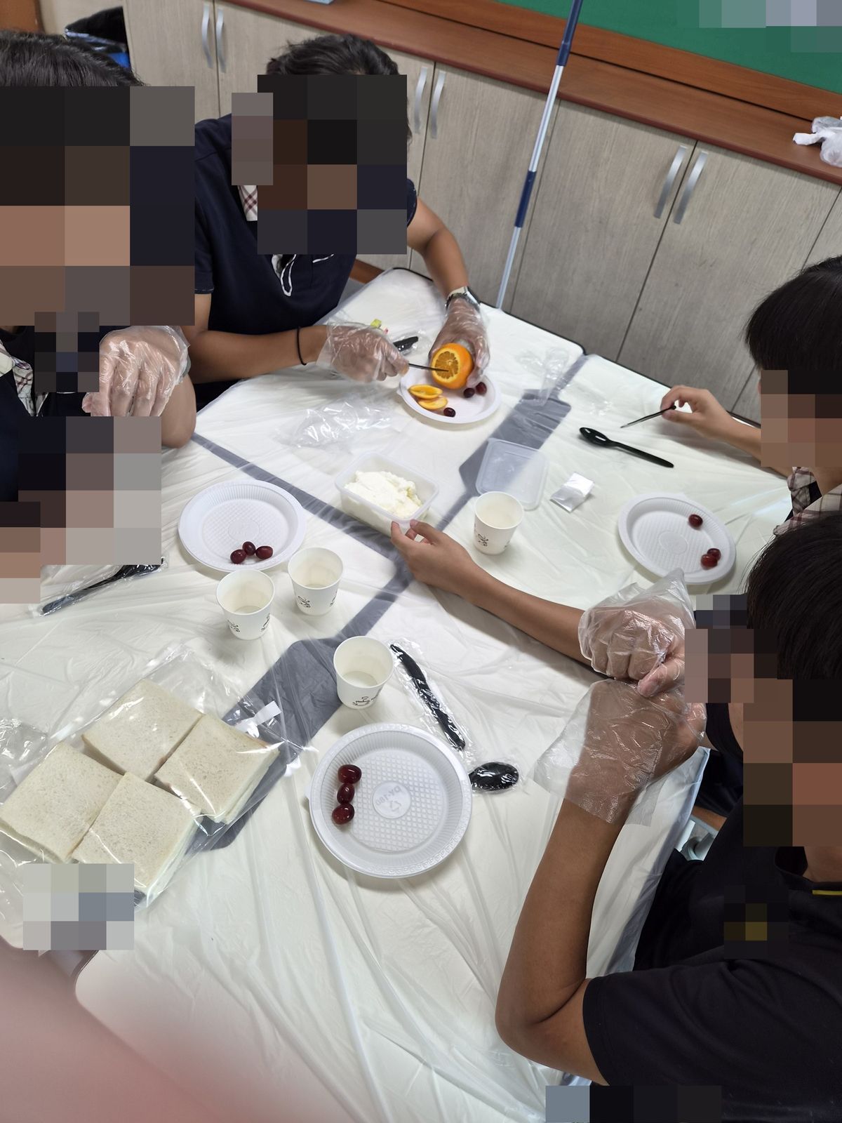 모둠 식탁에 네 명이 둘러앉아 식빵과 과일 접시, 크림 컵을 앞에 두고 장갑 낀 손으로 재료를 손질하는 모습