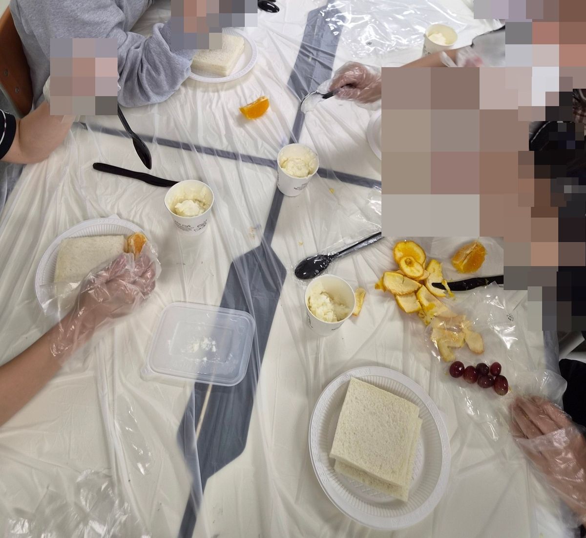 비닐을 덮은 식탁 위 식빵 접시와 크림 컵, 껍질 벗긴 과일 조각 사이로 장갑 낀 손들이 오가는 모습