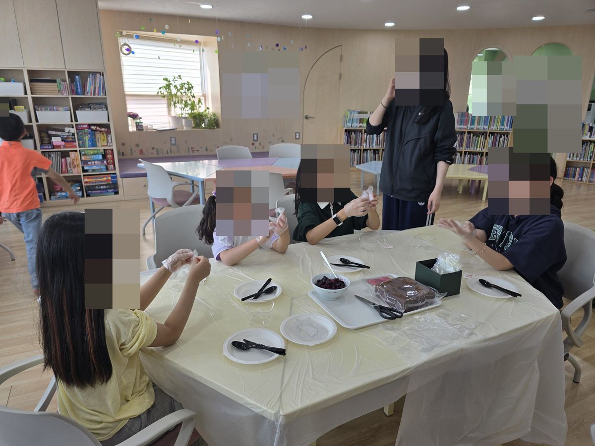 연노랑 식탁보를 깐 긴 테이블에 아이들이 둘러앉아 일회용 장갑을 끼고, 쟁반 위 비닐에 싸인 케이크와 베리 그릇, 가위가 놓인 모습