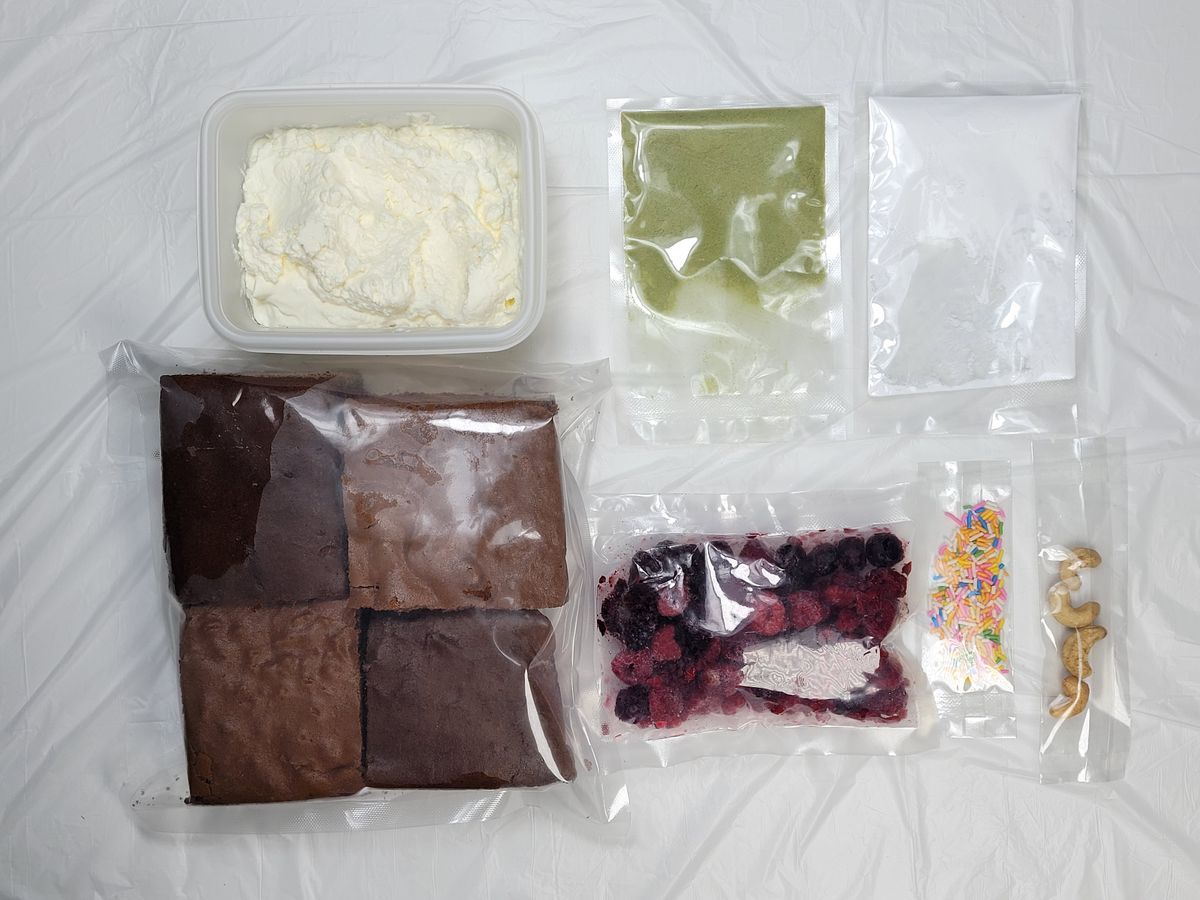 하얀 천 위에 갈색 케이크 시트 네 장과 하얀 크림 통, 과일 토핑·가루·장식 알갱이 봉지들이 한 벌로 펼쳐진 모습