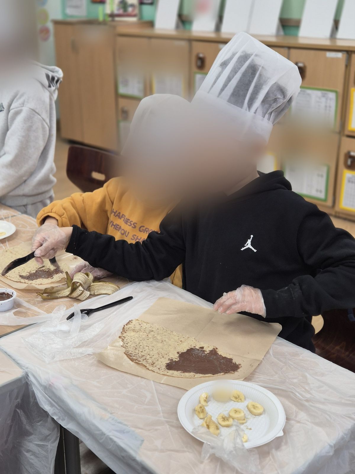 요리 모자를 쓰고 크레페빵에 스프레드를 바르는 아이의 손과 바나나 접시